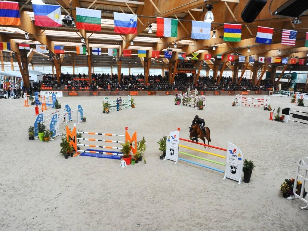 Manège équestre couvert avec cavaliers, obstacles colorés et drapeaux internationaux au-dessus de la tête.