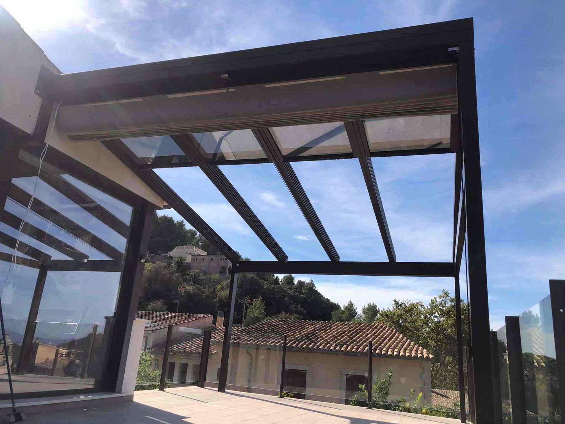 Una pérgola con techo de cristal y una casa al fondo.