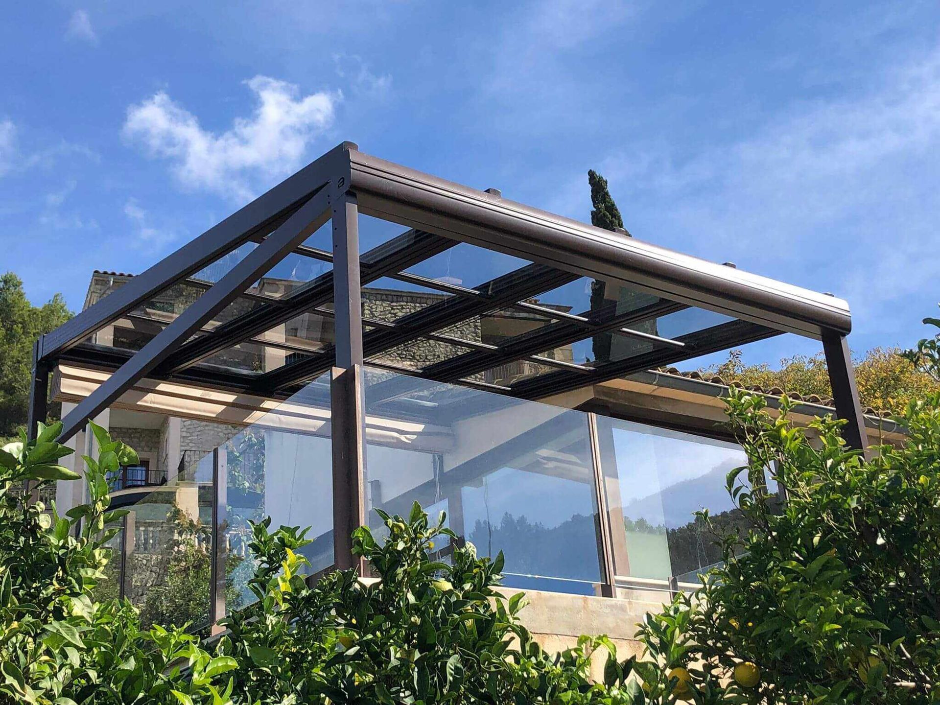 Una pérgola con techo de cristal está rodeada de árboles y arbustos.