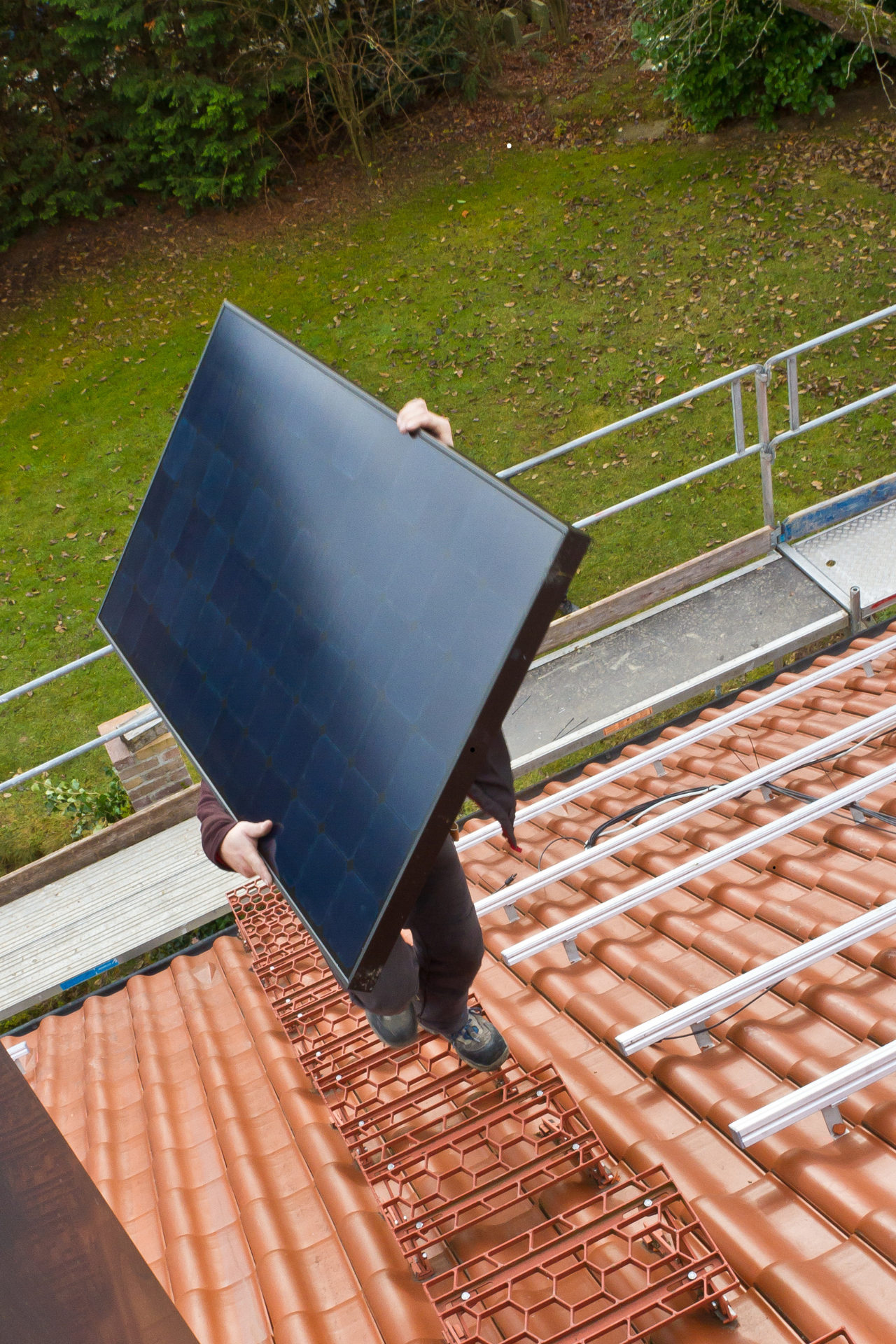 Personne installant un panneau solaire sur un toit de tuiles rouges, près d'une pelouse verte et d'un échafaudage.