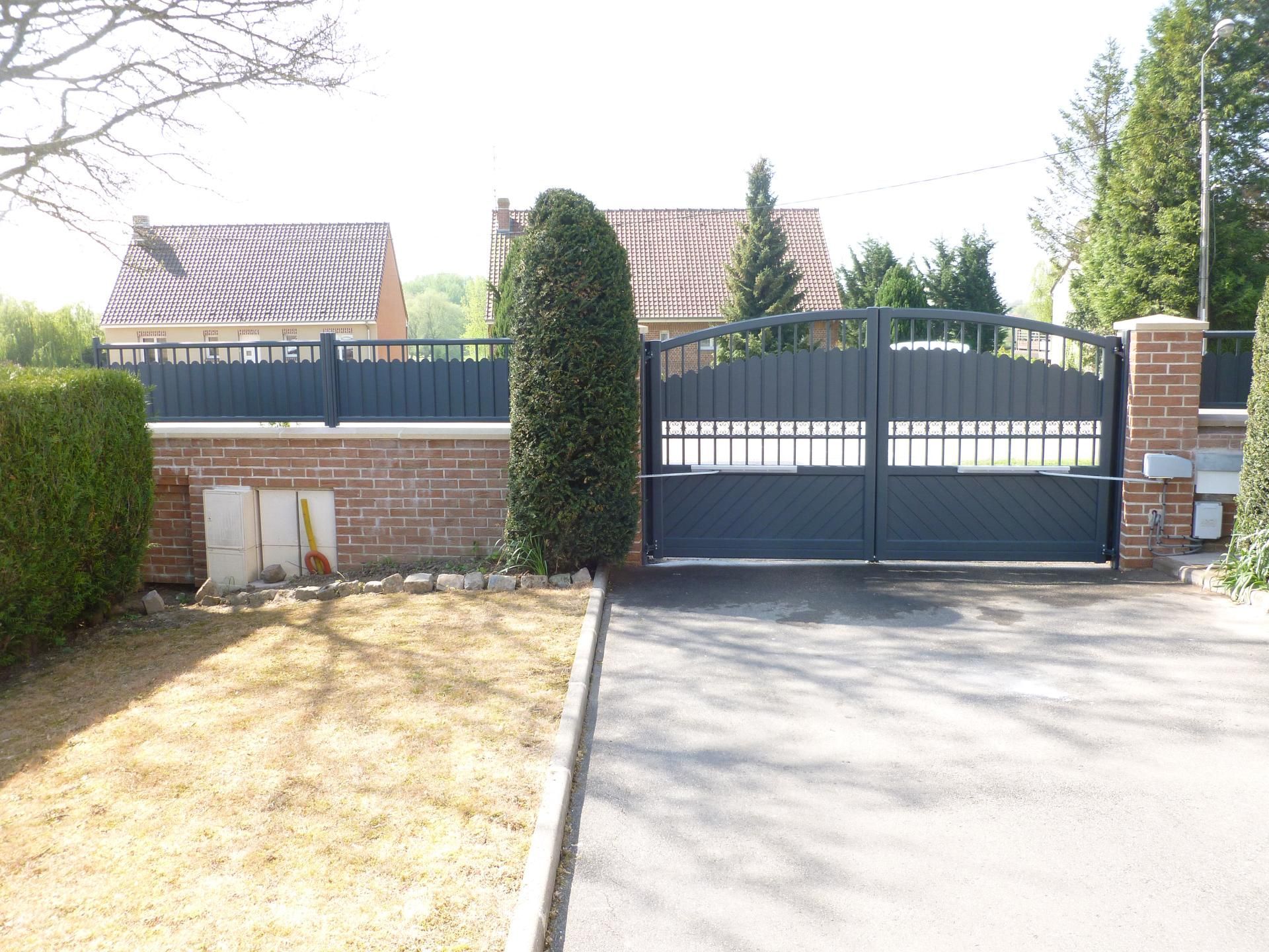 Portail et clôture noirs sur un mur de briques devant une maison, avec allée et cour.
