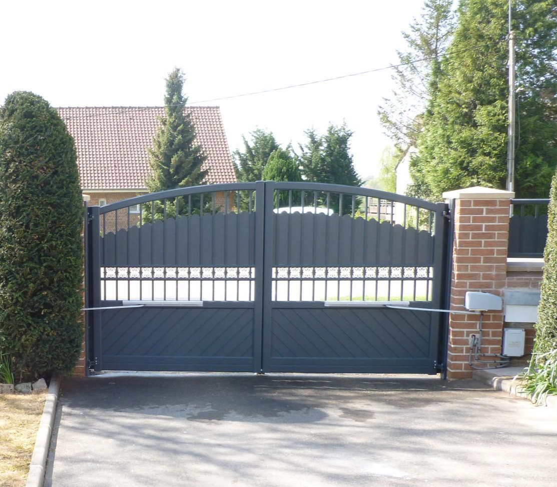 Portail d'allée en métal gris devant une maison. Deux portails sont fermés. Buissons verts à gauche et pilier en briques à droite.