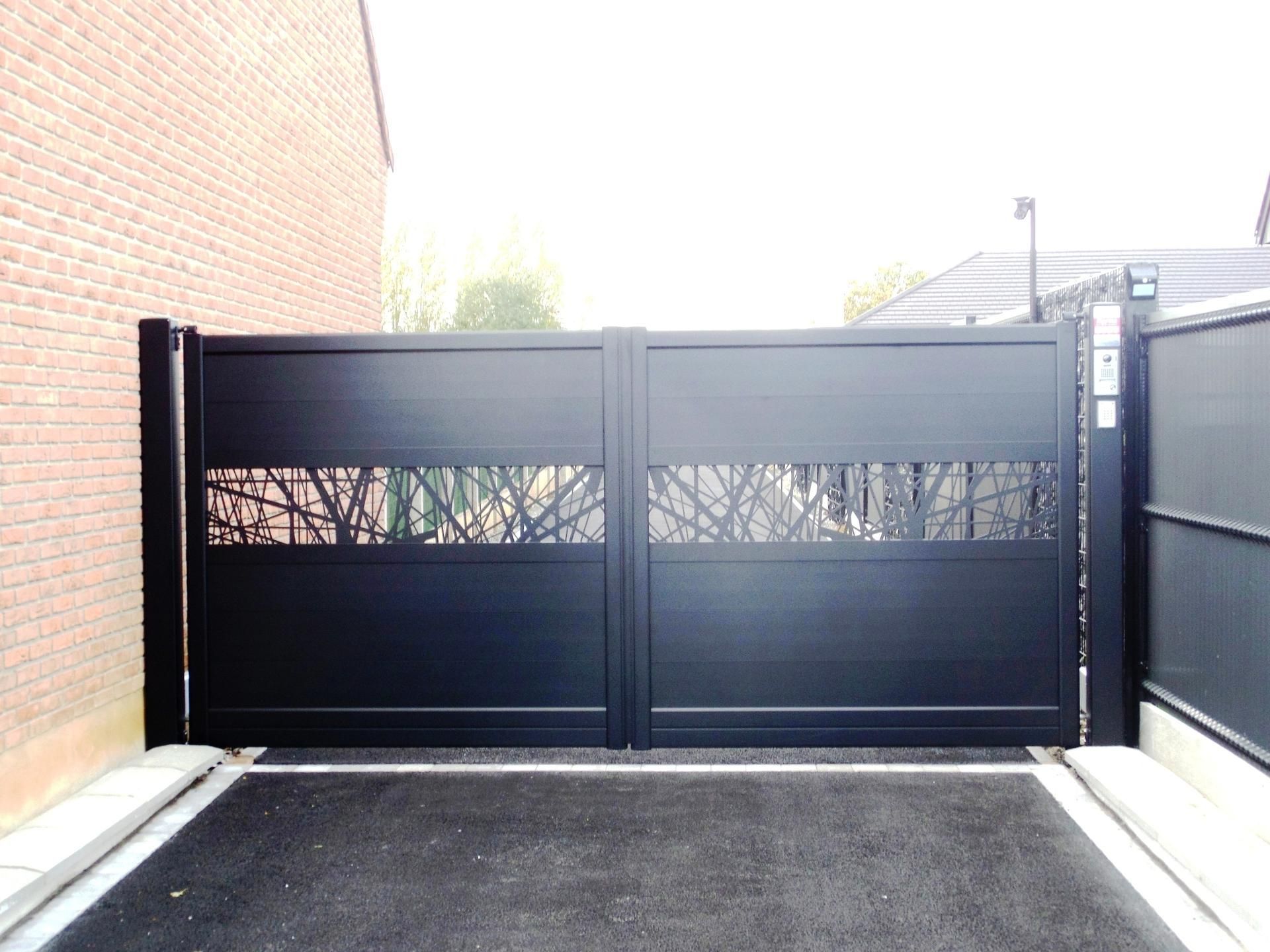 Portail d'allée en métal noir avec des découpes en forme d'arbre, sur une surface pavée, à côté d'un bâtiment en briques.