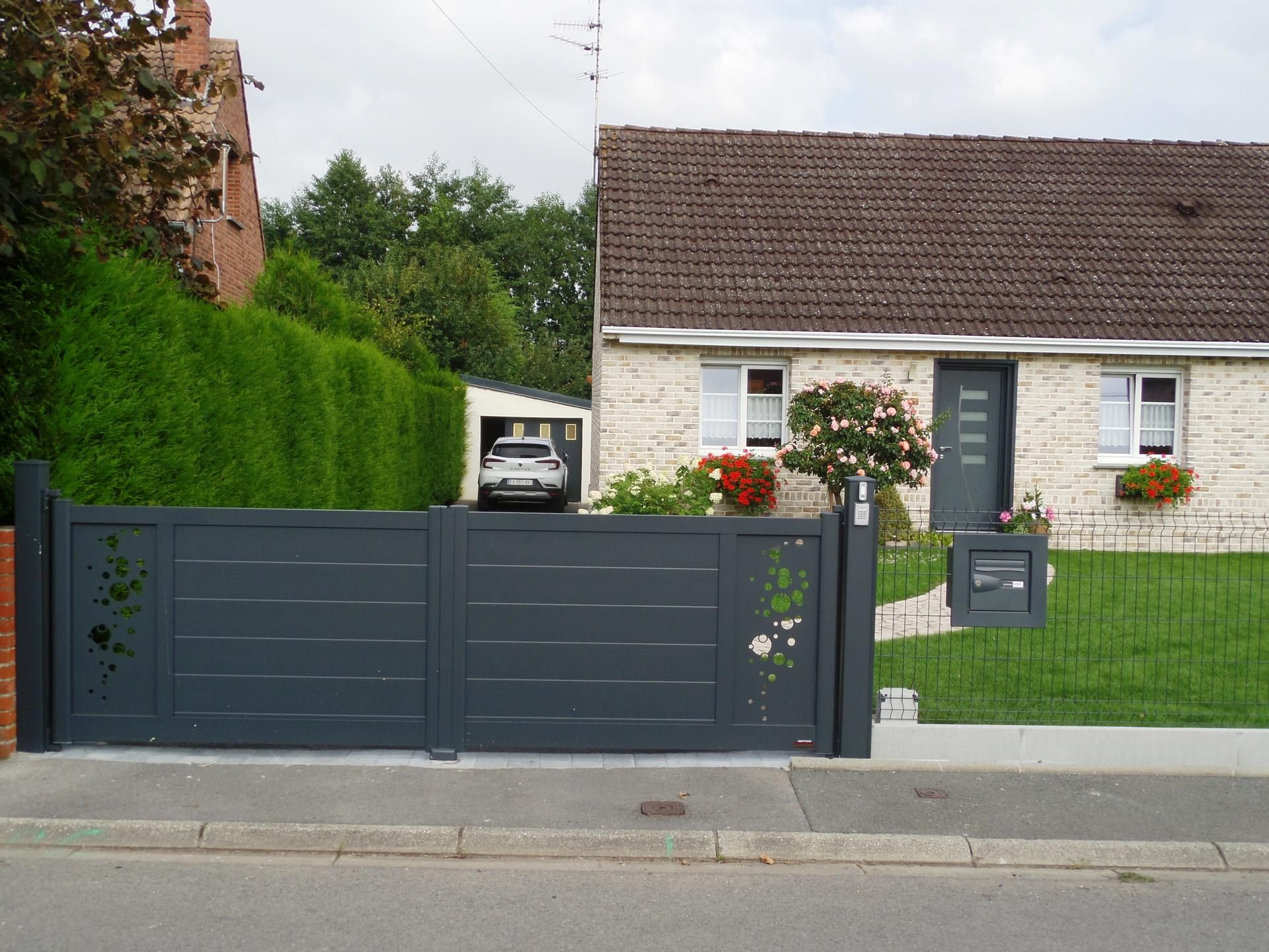 Maison grise avec portail assorti, voiture garée dans le garage, pelouse verte, arbres et ciel nuageux.