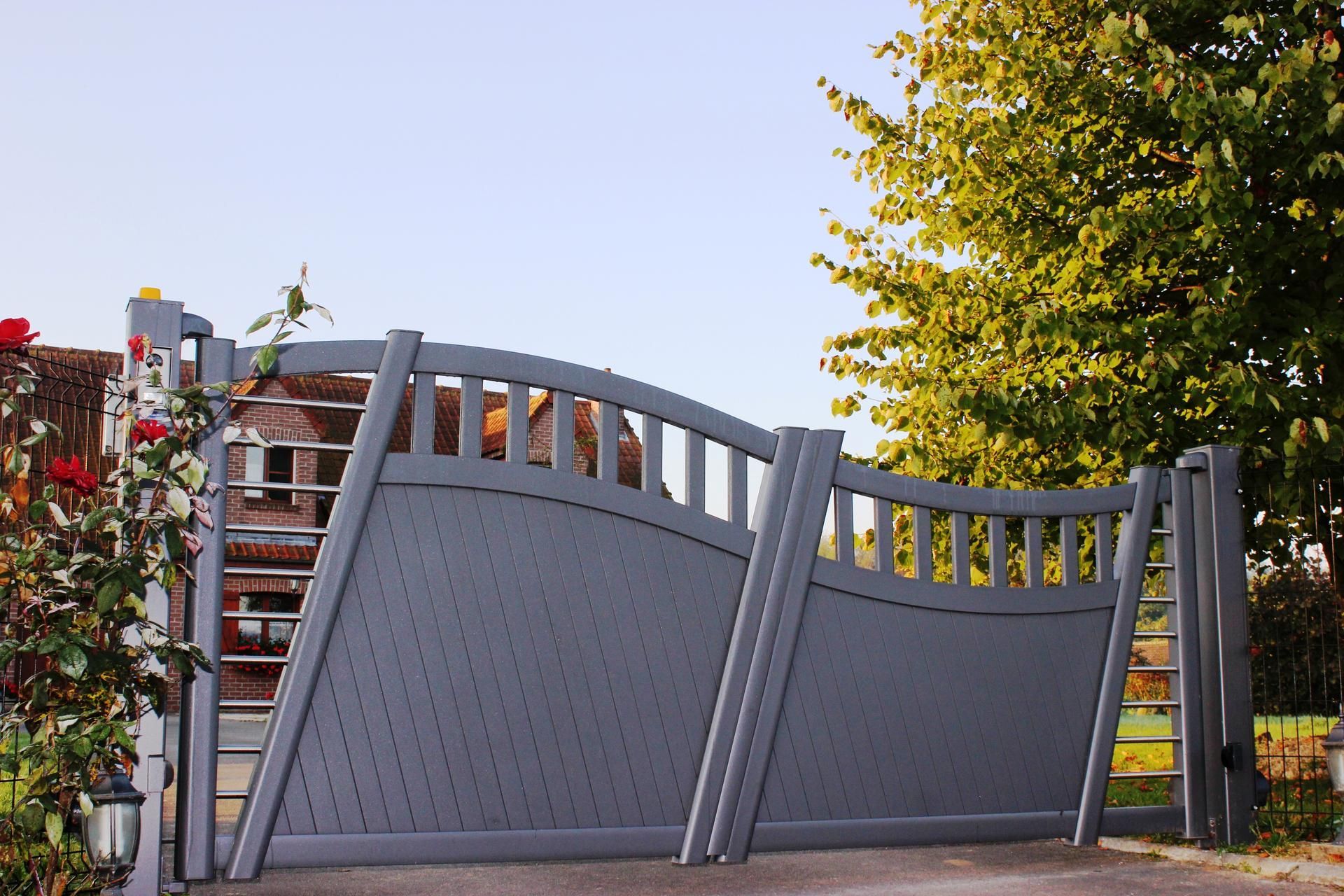 Portail d'allée gris et incurvé avec un ornement décoratif, devant une maison et des arbres.