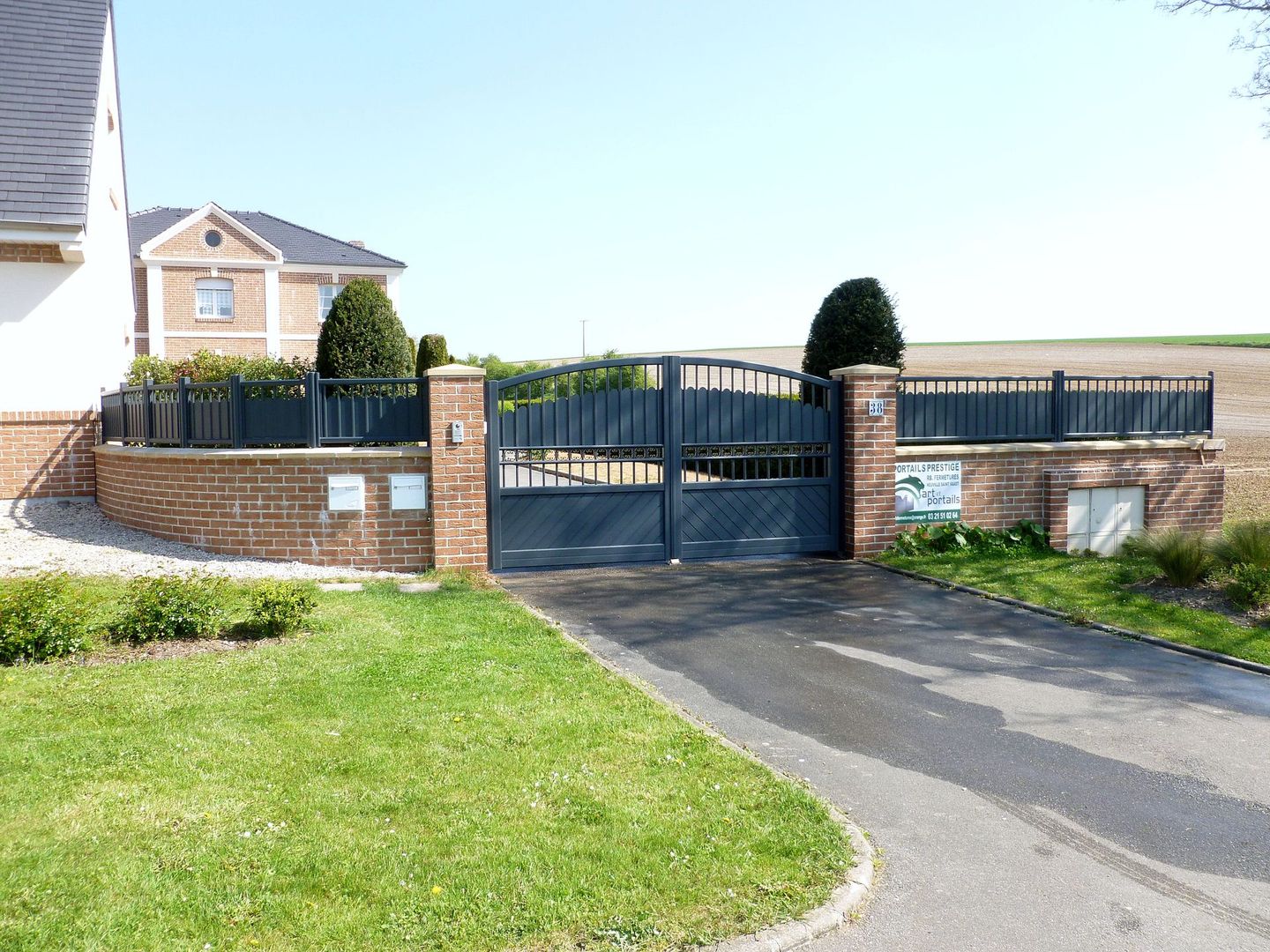 Portail d'allée gris et incurvé avec un ornement décoratif, devant une maison et de la verdure.