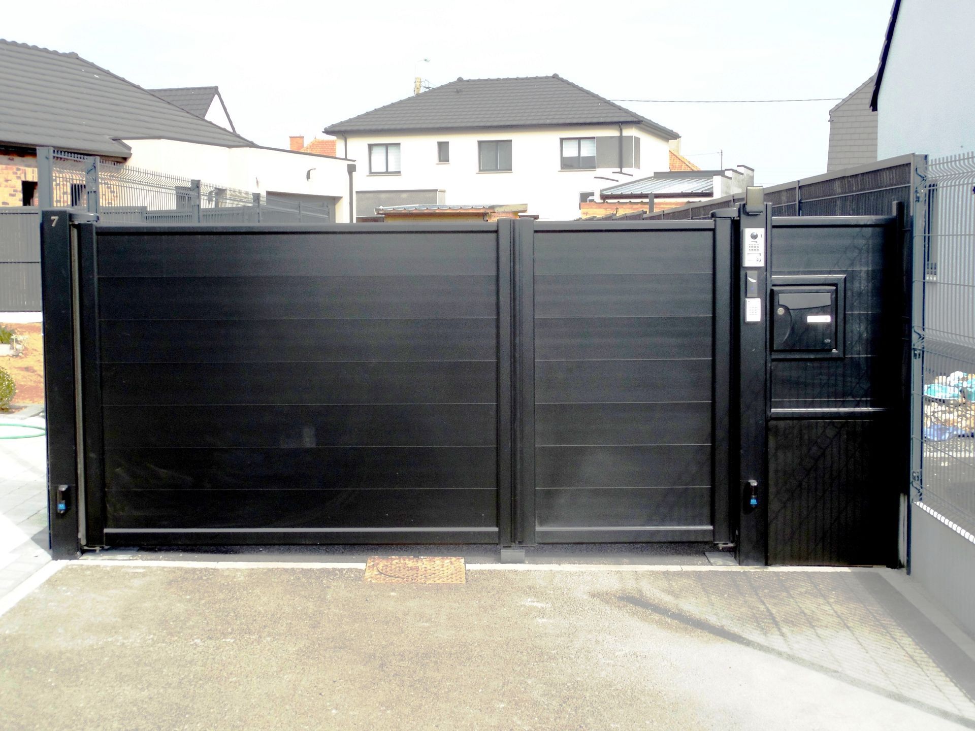 Un portail et une clôture en métal gris foncé, sur un mur de briques, mènent à une maison au toit de tuiles.