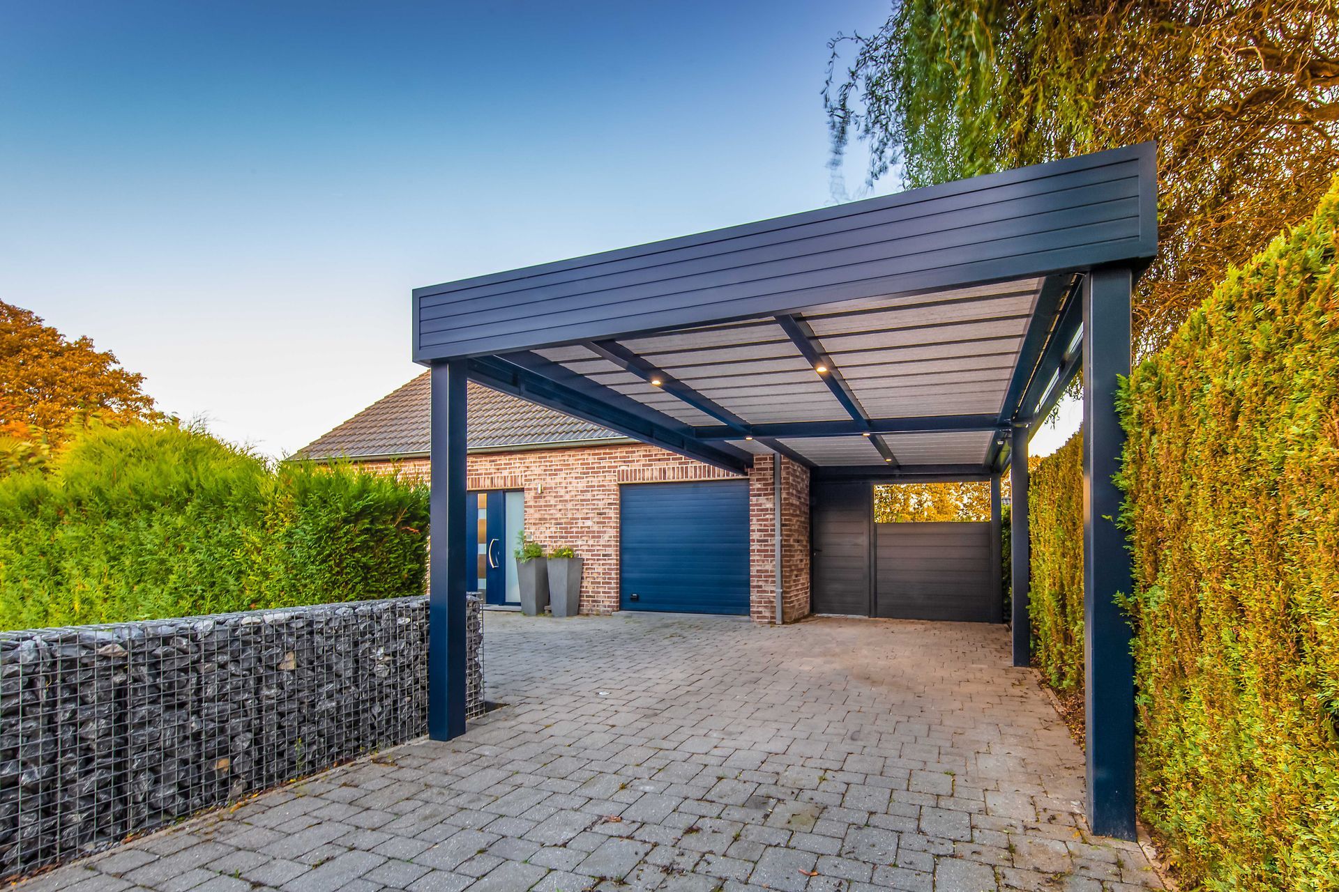 Pergola à structure grise et toit transparent, jouxtant une maison avec une pelouse verte.