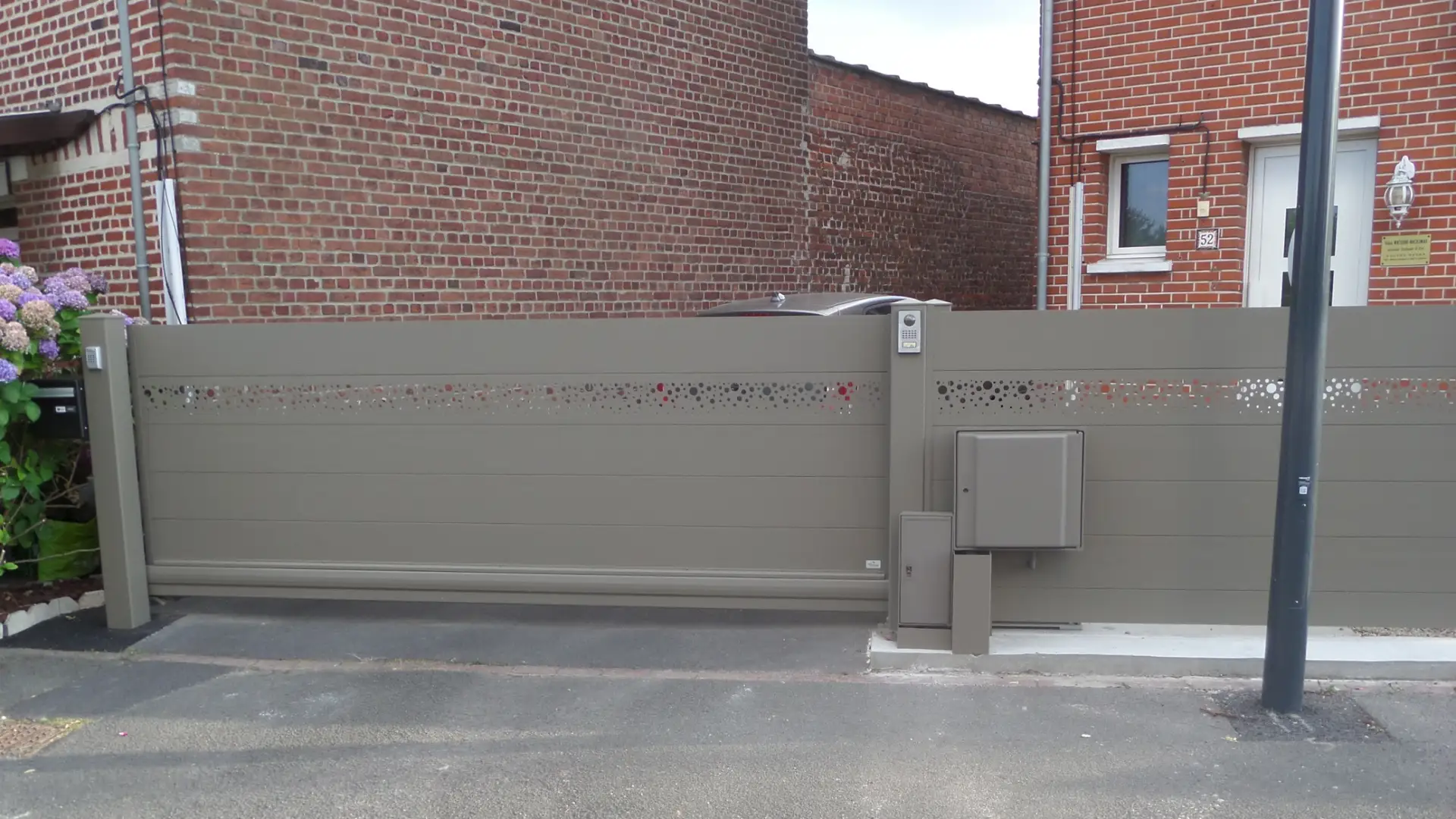 Portail coulissant beige orné de découpes décoratives, devant un bâtiment en briques. On aperçoit la rue et un lampadaire.