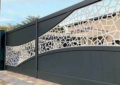 Portail en métal gris foncé orné de découpes abstraites blanches ; se détachant sur un bâtiment et un ciel de couleur claire.