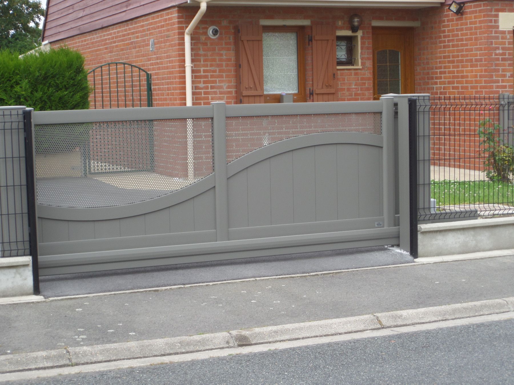 Portail coulissant gris au design incurvé, devant une maison en briques avec une allée pavée.
