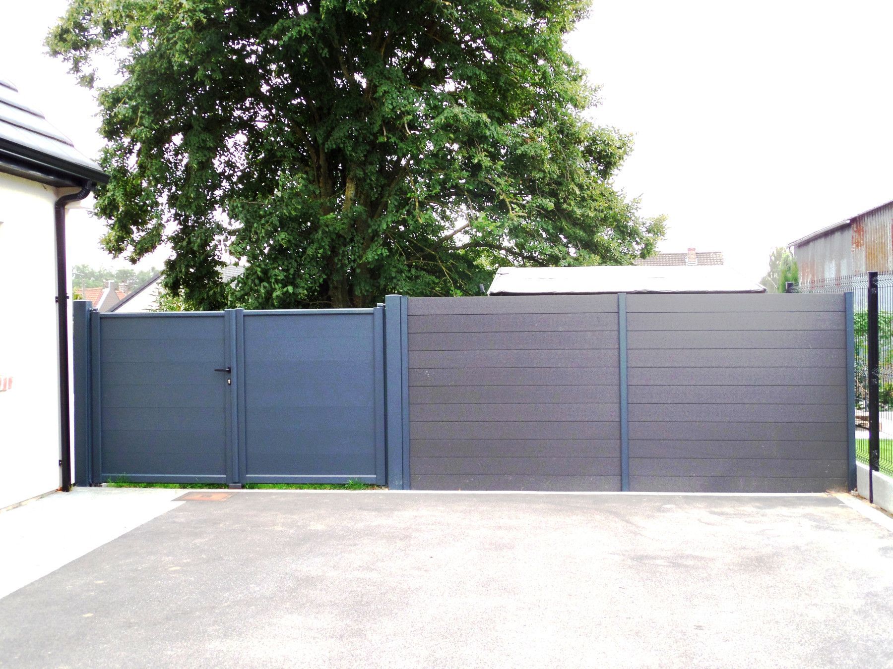 Portail et clôture gris devant une maison, avec un arbre en arrière-plan.