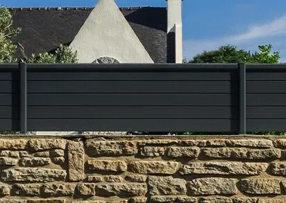 Clôture à lattes horizontales gris foncé sur un mur de pierre patiné ; bâtiment et ciel en arrière-plan.