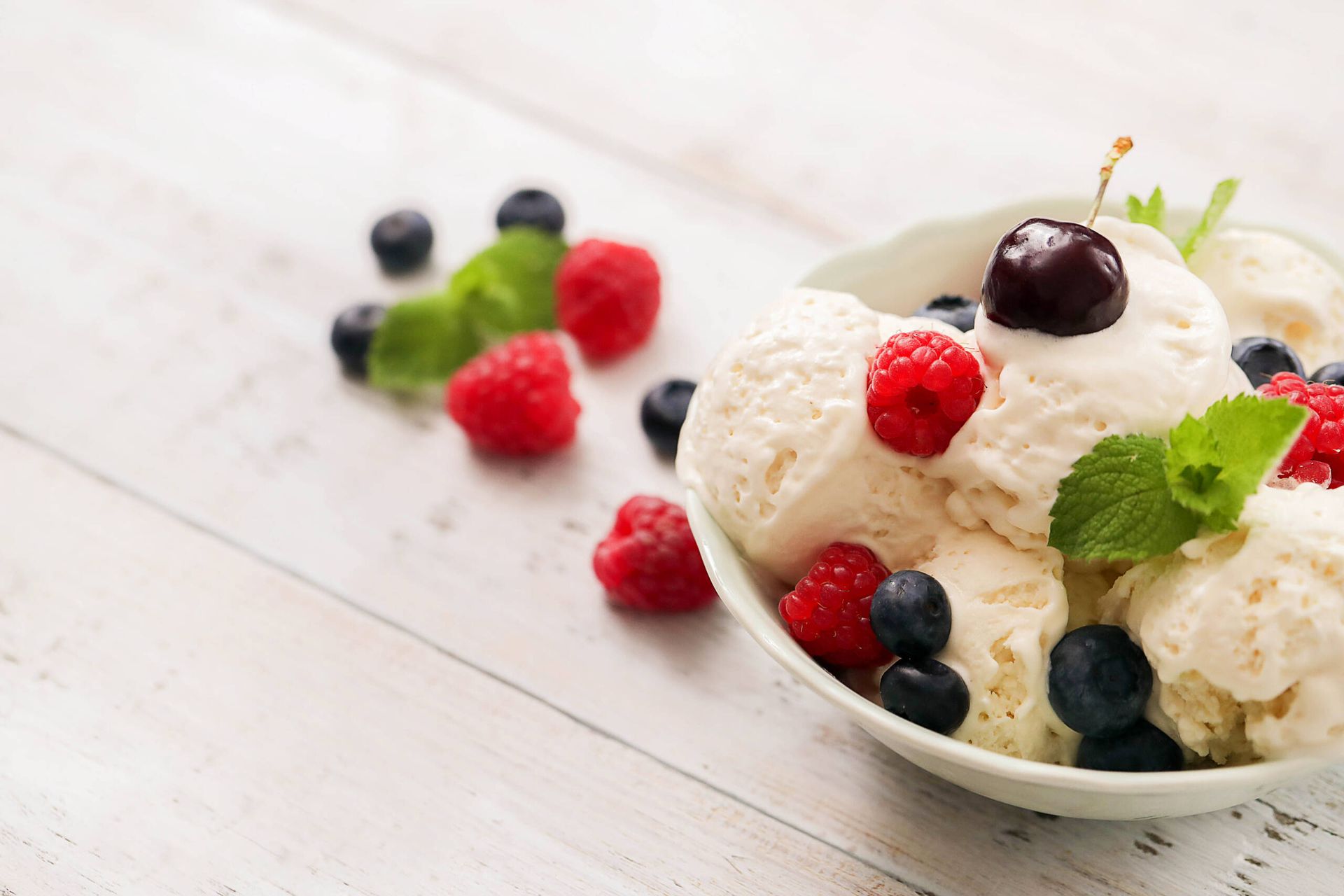 Helado de vainilla en un cuenco, cubierto con frutos rojos y menta, sobre una mesa de madera blanca