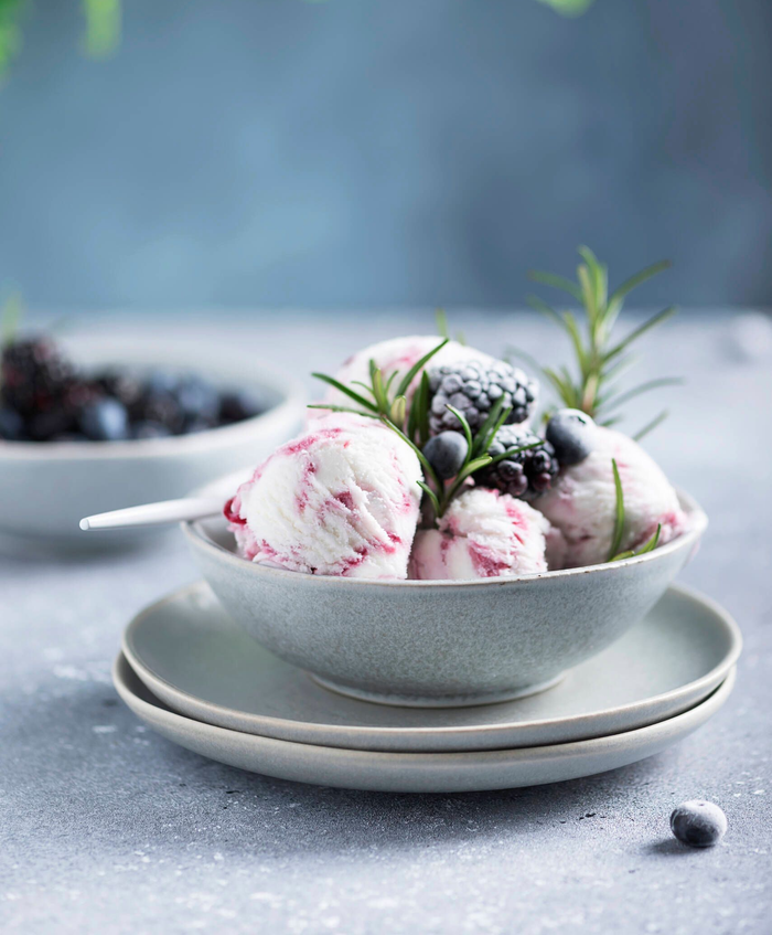 Cuenco con bolas de helado de frutos rojos y menta por encima, sobre platos apilados y con frutos rojos desenfocados al fondo