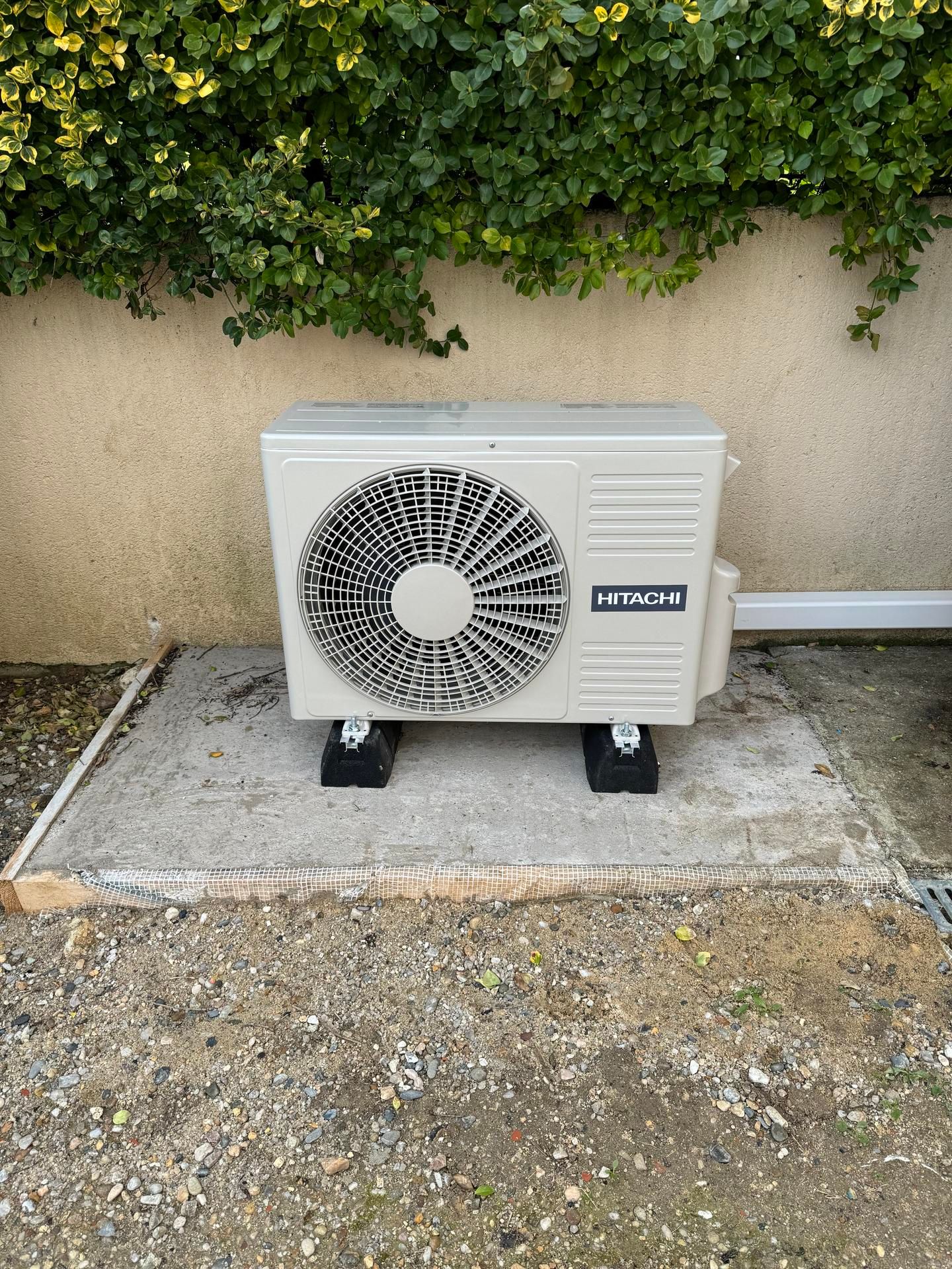 Unité de pompe à chaleur sur dalle de béton, à côté d'un mur bordé de végétation.