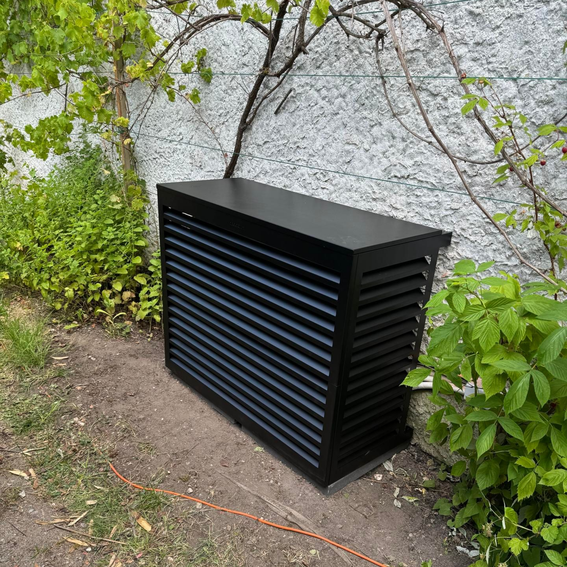 Cache-climatiseur noir à lamelles horizontales sur un mur blanc texturé, entouré de feuillage et d'herbe.