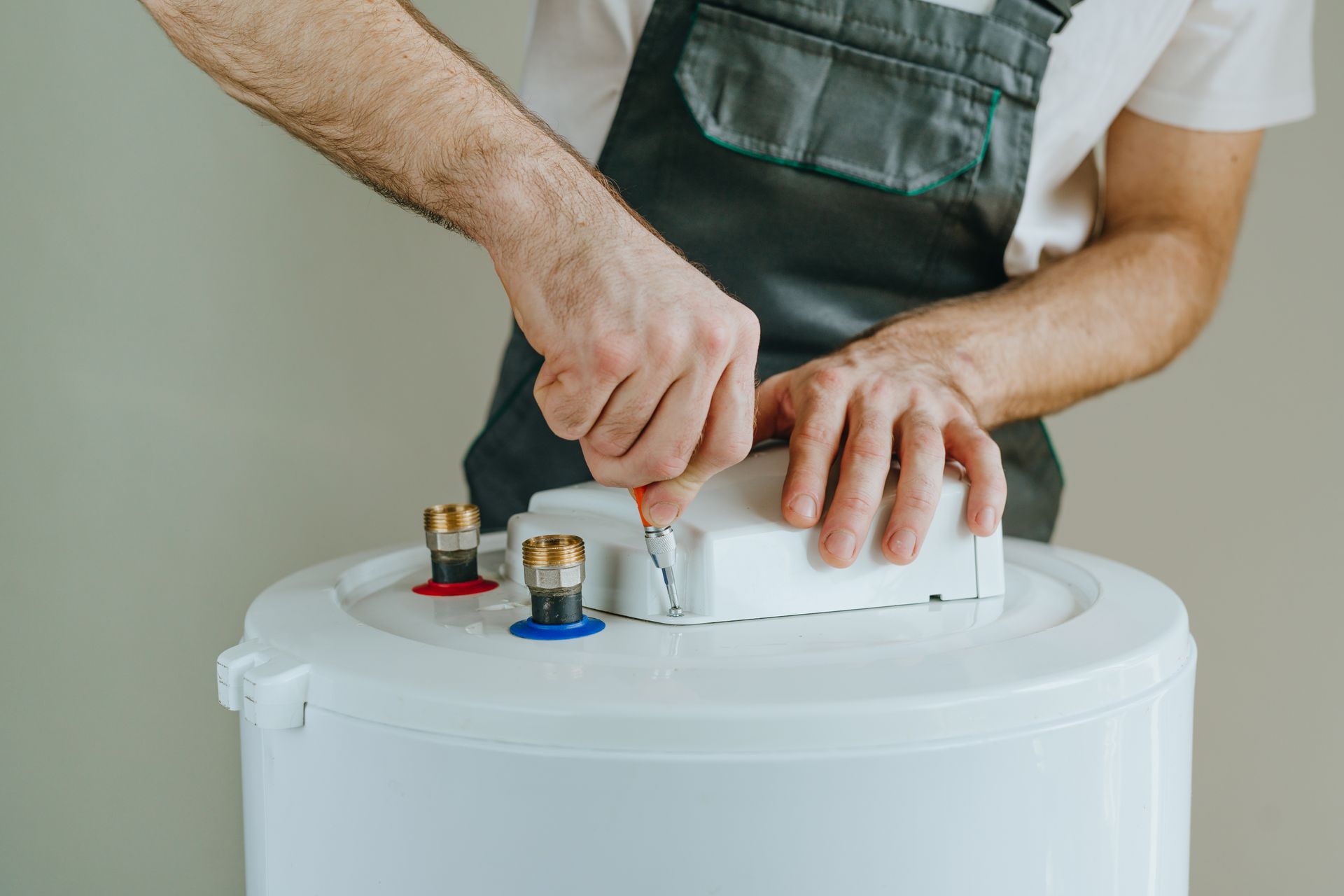 Une personne en salopette utilise un tournevis sur un chauffe-eau blanc.