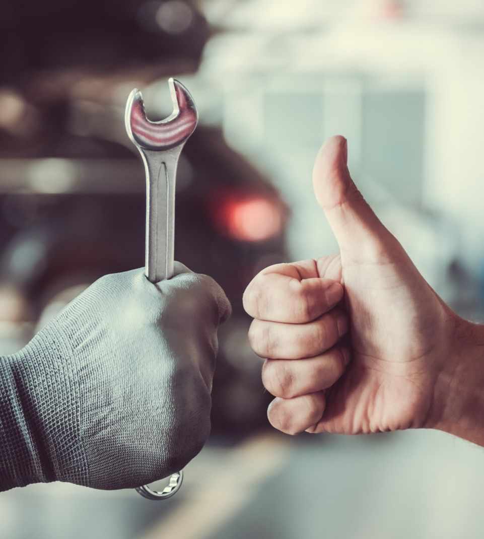 Dans un atelier automobile, une main gantée tient une clé à molette tandis qu'une main nue fait un signe avec le pouce.
