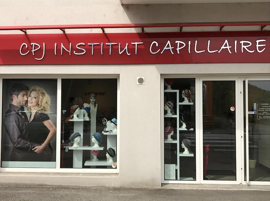 Extérieur d'un institut de coiffure avec des mannequins portant des perruques exposés en vitrine. Un auvent rouge porte l'inscription « CPJ INSTITUT CAPILLAIRE ».