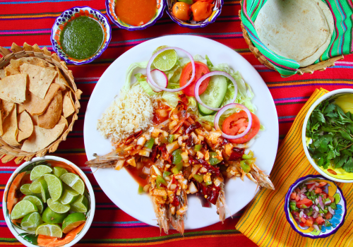 Mexican dish: Fried whole fish with salsa, rice, salad, tortilla chips, limes, salsas, and tortillas on a colorful table.