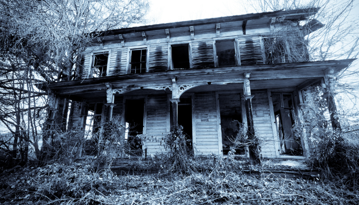 Dilapidated, two-story house with missing windows and overgrown vegetation, ominous and gray.