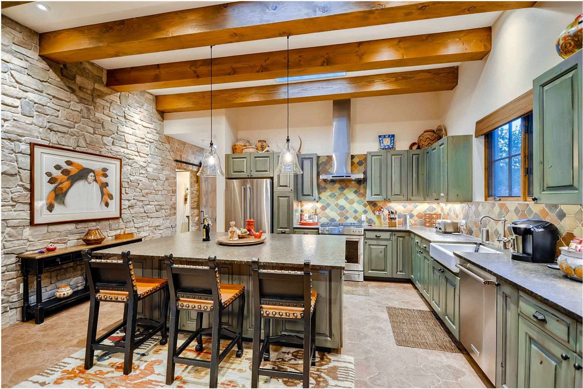 Rustic kitchen with stone wall, green cabinets, island with stools, and wood beams.