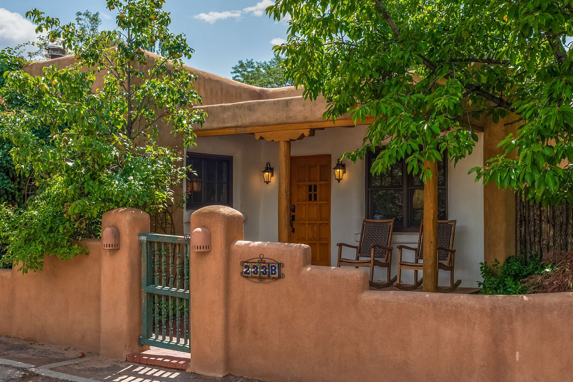 Adobe-style home with stucco walls, a green gate, and trees, with a brown wooden door and two rocking chairs.