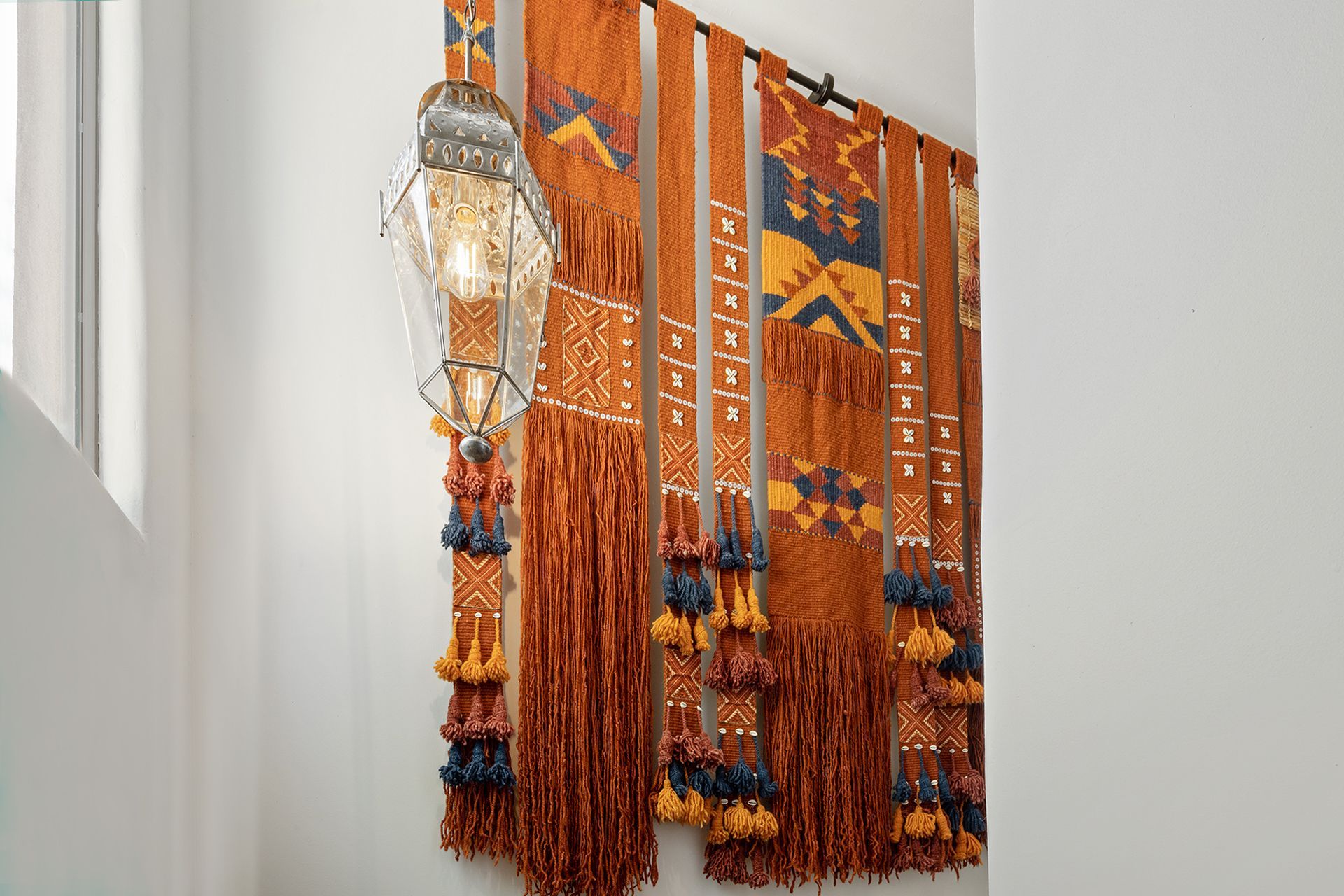 Textile wall hanging in orange, yellow, and blue with a decorative light fixture to the left.