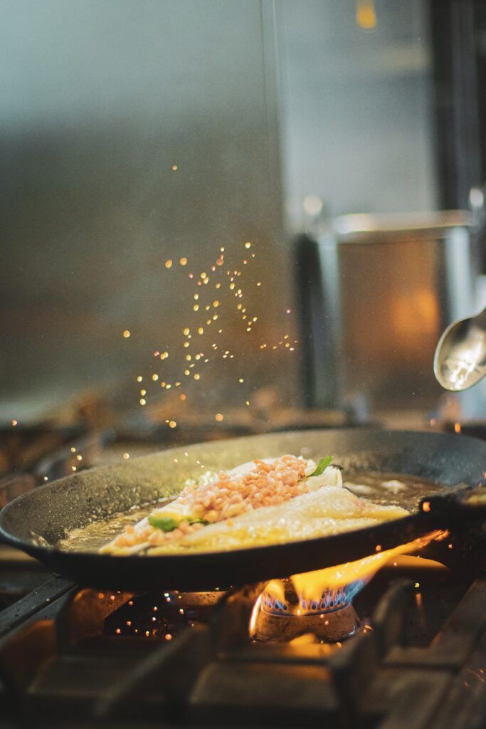 Ein Koch brät Fisch in einer Pfanne über einer Gasflamme, Funken sprühen in der Küche.