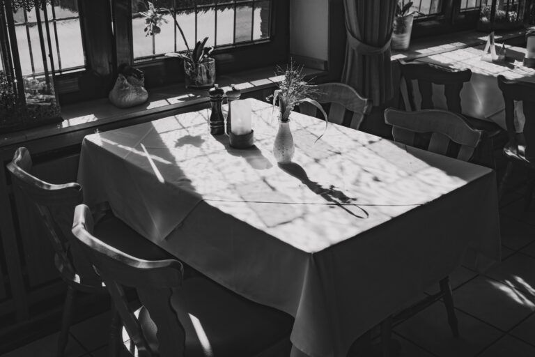 Ein sonnenbeschienener Esstisch am Fenster mit einer Vase, einer Flasche und Gläsern, die lange Schatten werfen.
