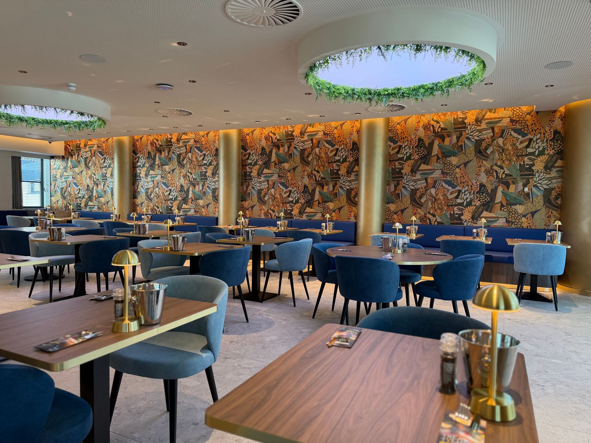 Moderne restaurantzaal met blauwe stoelen, houten tafels en een muur met een patroon en groene ronde plafondlampen.
