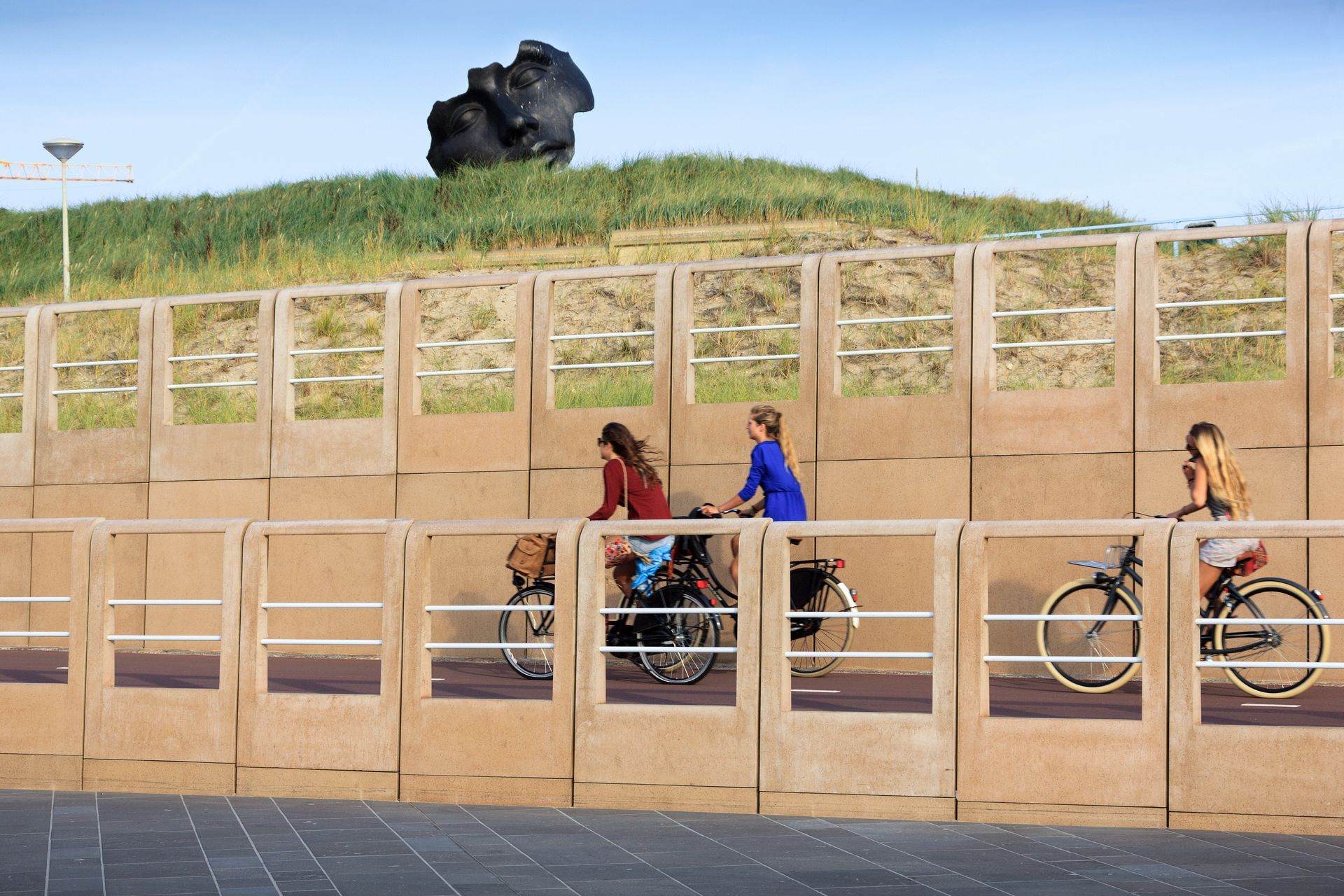 Mensen wandelen en fietsen langs een houten hek onder een grasheuvel met een sculptuur erop.