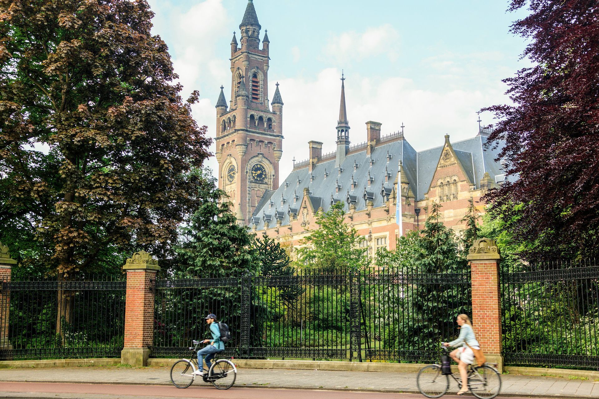 Fietsers passeren een smeedijzeren hek met daarachter een historische klokkentoren en een bakstenen gebouw.