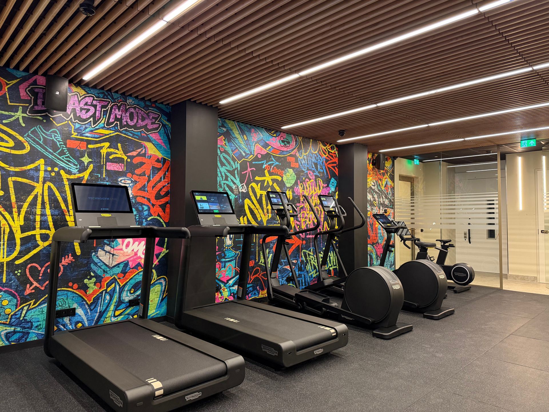 Fitnessruimte met loopbanden, crosstrainers, muren met graffiti-schilderingen en plafondverlichting met houten latten.