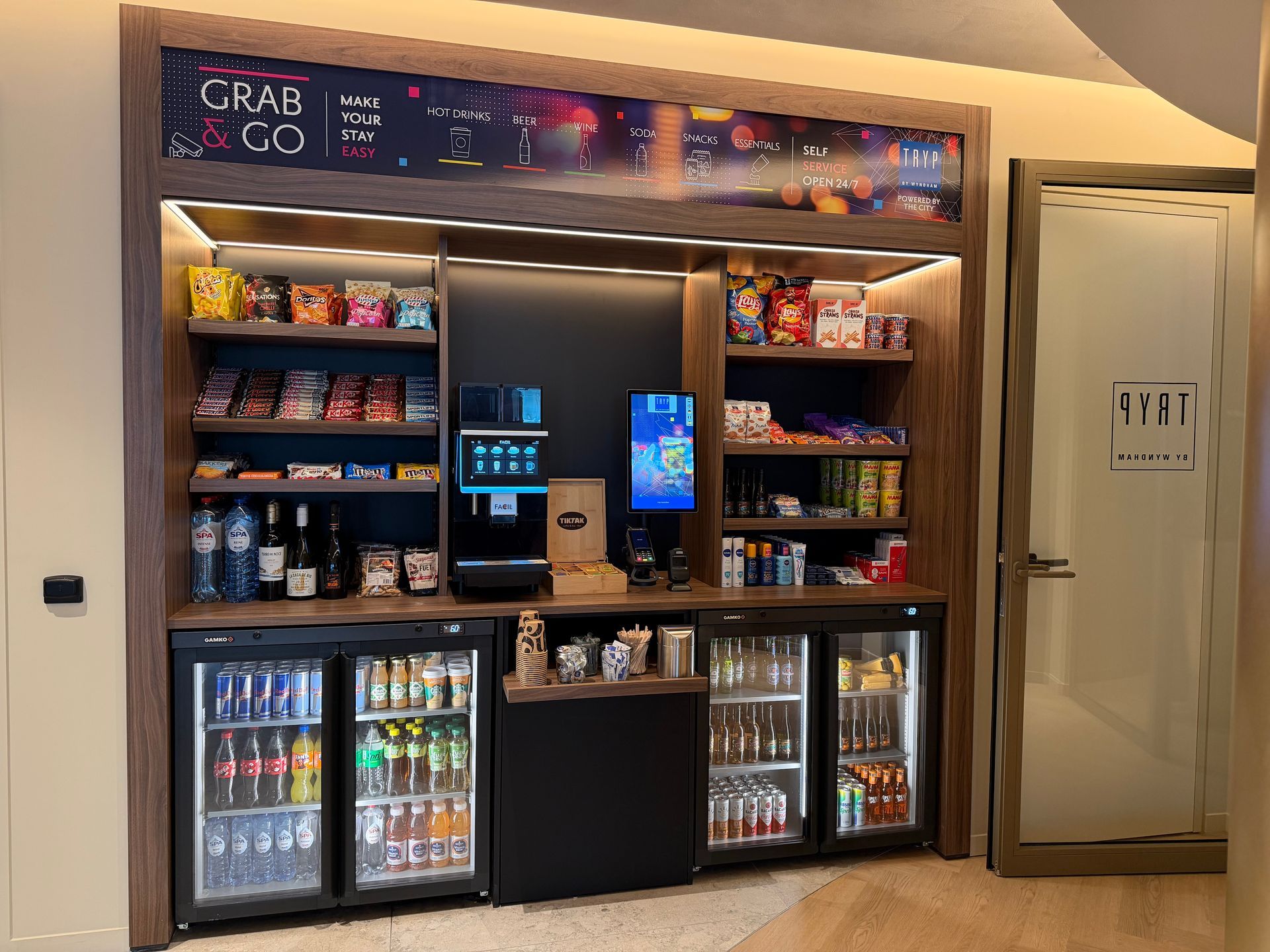 Kleine bar met verlichte drankkoelers, schappen vol snacks en kleurrijke menukaarten.