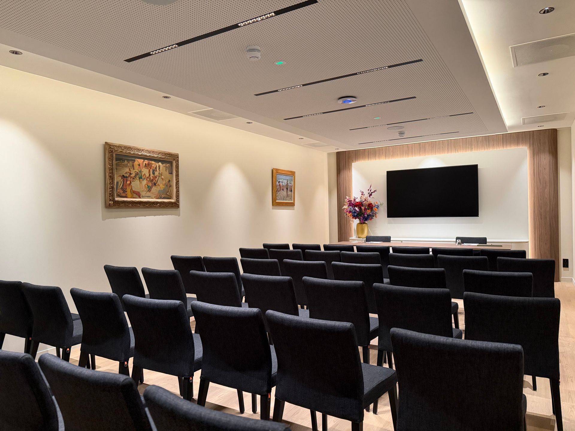 Vergaderruimte met rijen zwarte stoelen tegenover een aan de muur gemonteerde tv en een podium, zacht verlicht.