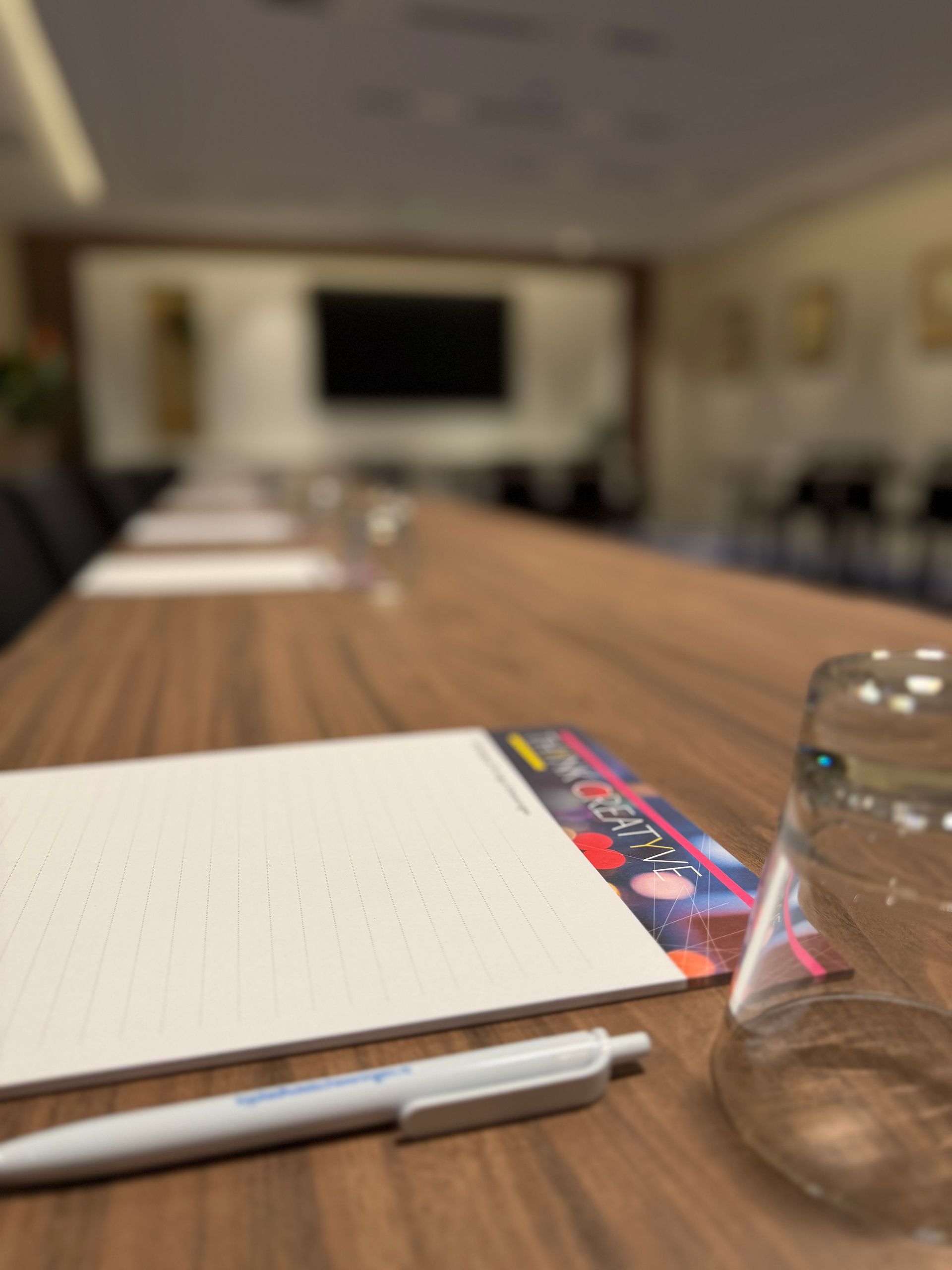 Notitieboekje en pen op een vergadertafel, met een wazige vergaderruimte en presentatiescherm op de achtergrond.