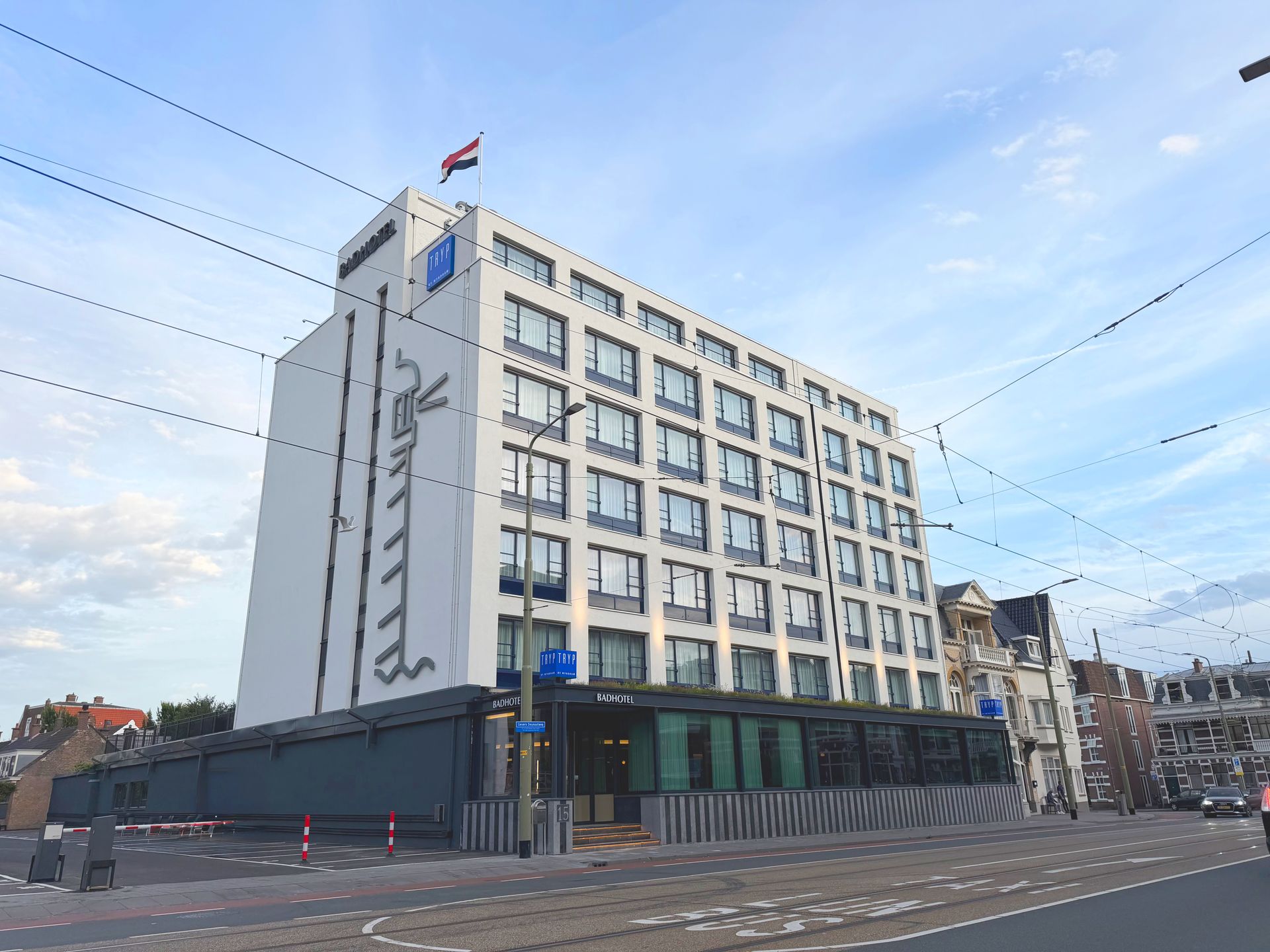 Modern kantoorgebouw met meerdere verdiepingen, een witte gevel, een glazen begane grond en vlaggen op het dak onder een blauwe hemel.