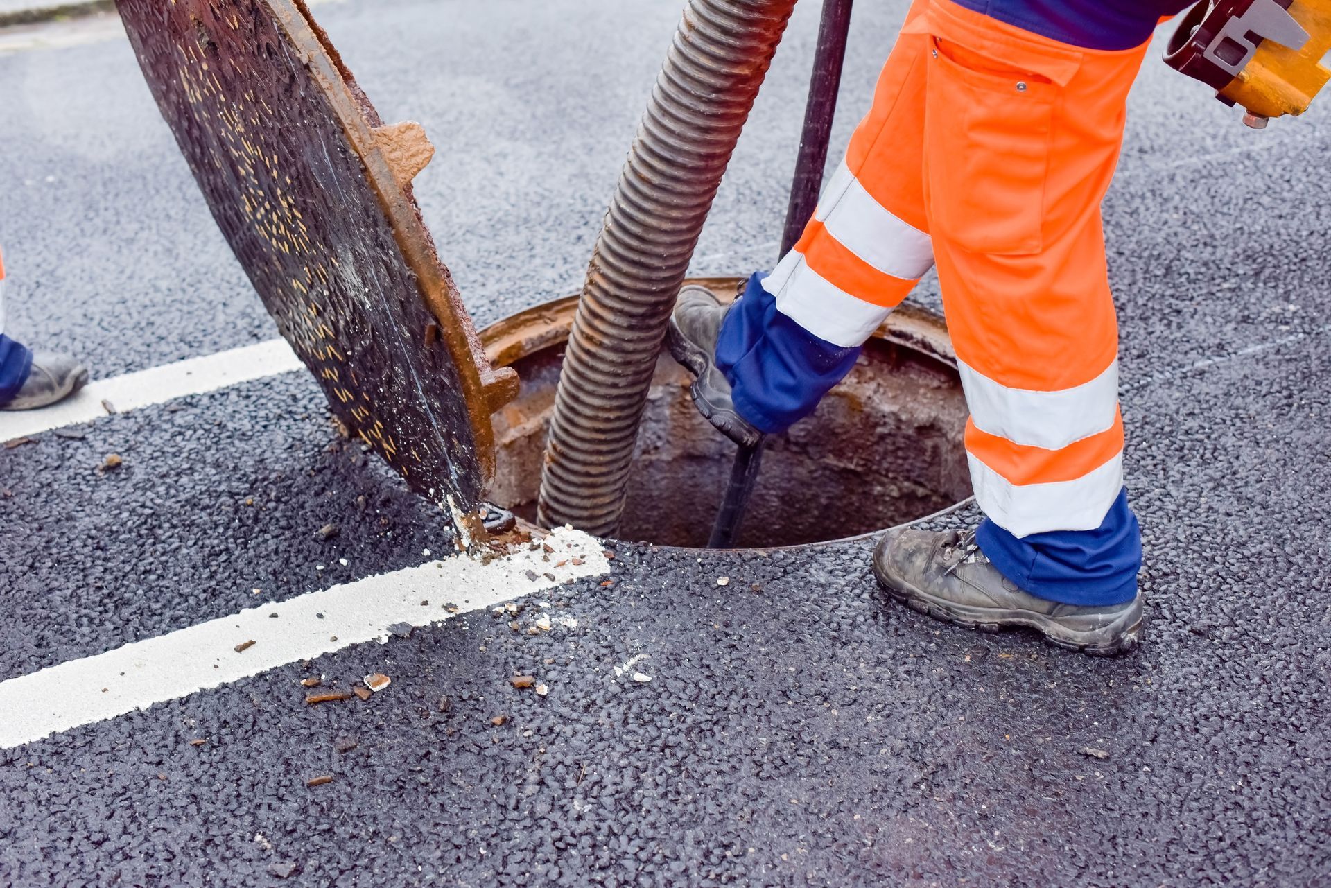 Personne en pantalon de travail orange et bottes nettoyant une plaque d'égout sur une route pavée.