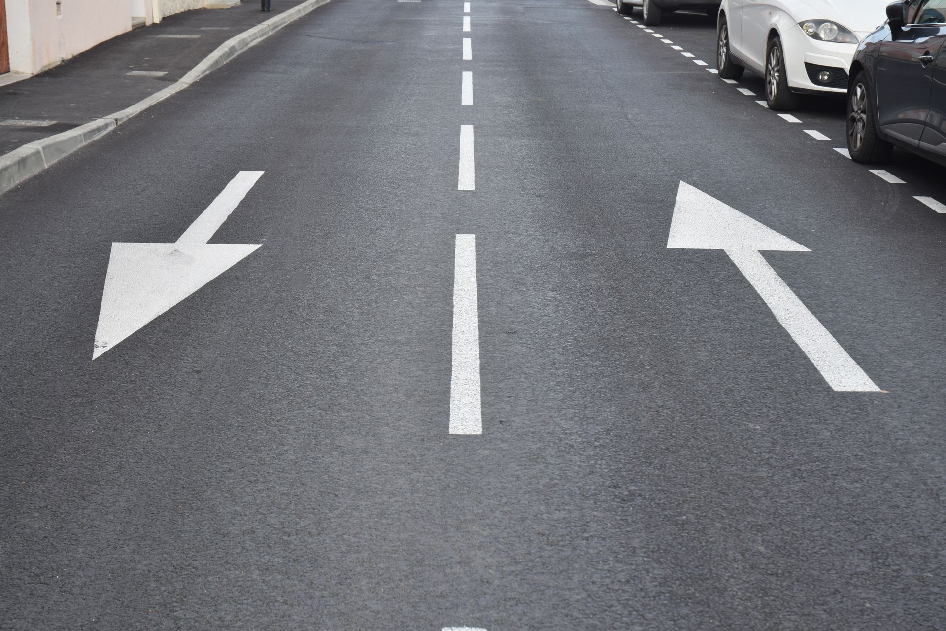 Route avec flèches directionnelles blanches : gauche, droite et tout droit. Voitures stationnées à droite.