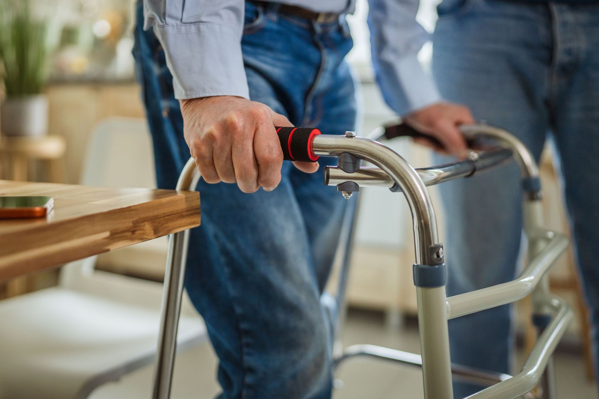 Une main tenant un déambulateur à poignée rouge, à côté d’une autre personne en jean, à l’intérieur