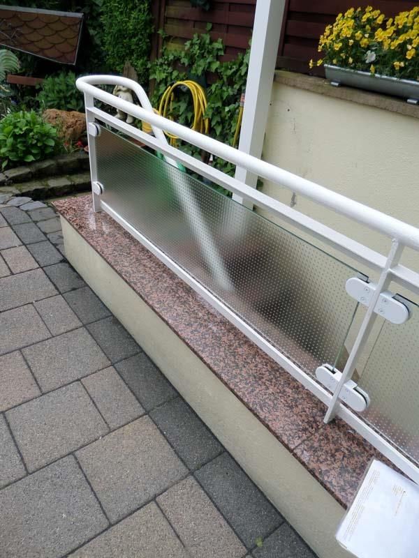 Ein weißes Geländer mit einer klaren Glasscheibe auf einem gemauerten Gehweg