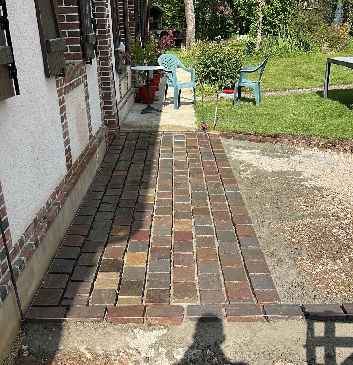 Allée en briques longeant une maison et menant à une cour gazonnée avec deux chaises de jardin et une petite table.