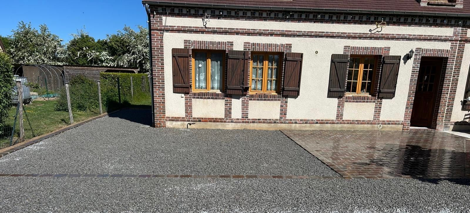 Cour de gravier jouxtant une maison blanche aux volets bruns et à la porte en bois, sous un ciel bleu.