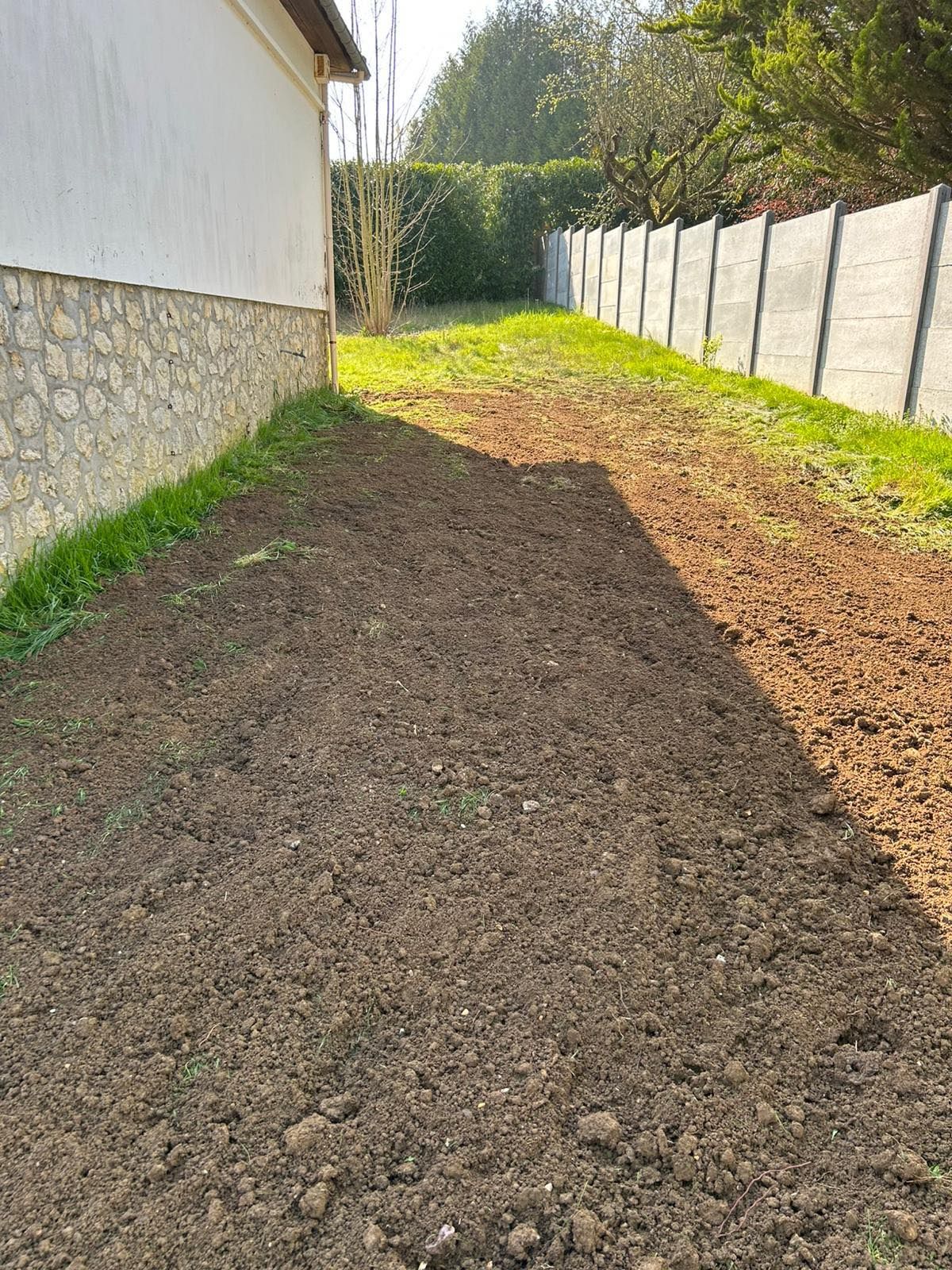 Bande de terre fraîchement labourée à côté d'une maison, bordée d'une clôture et de fondations en pierre dans une cour ensoleillée