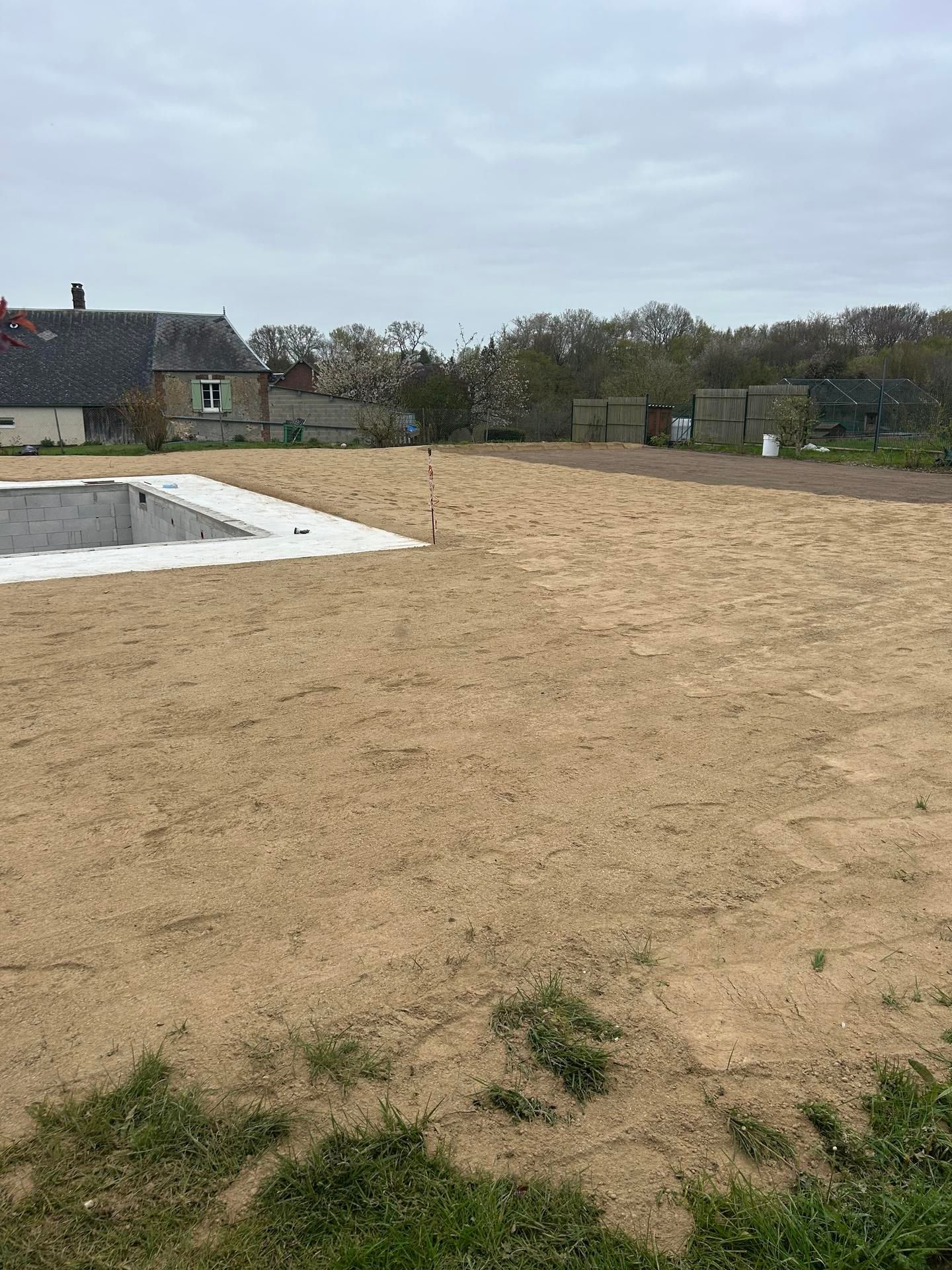 Terrain vague sablonneux jouxtant une maison, avec une fondation rectangulaire en béton au premier plan.