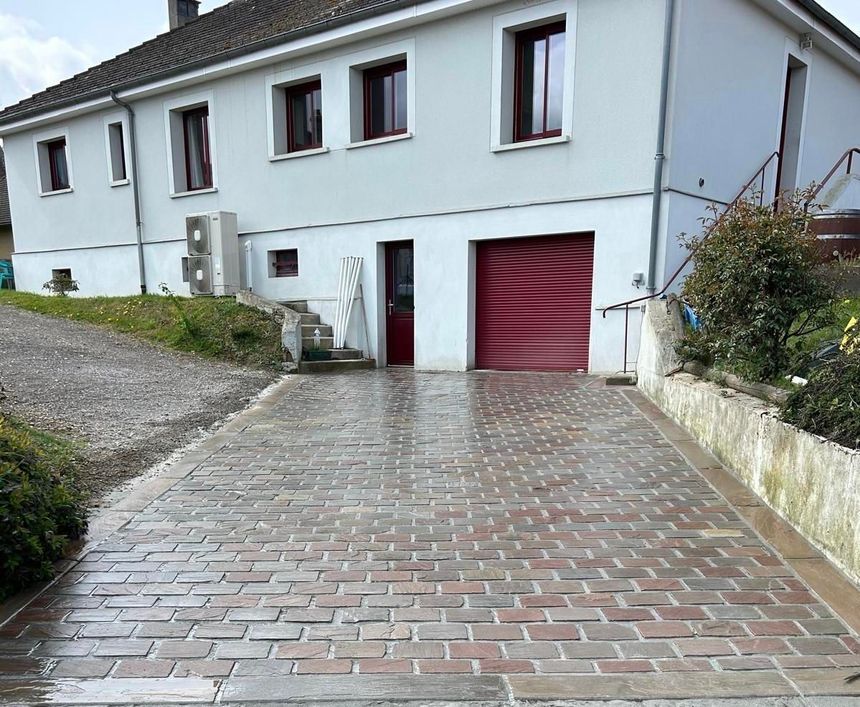 Maison blanche avec allée en briques et porte de garage rouge dans une rue résidentielle en pente.