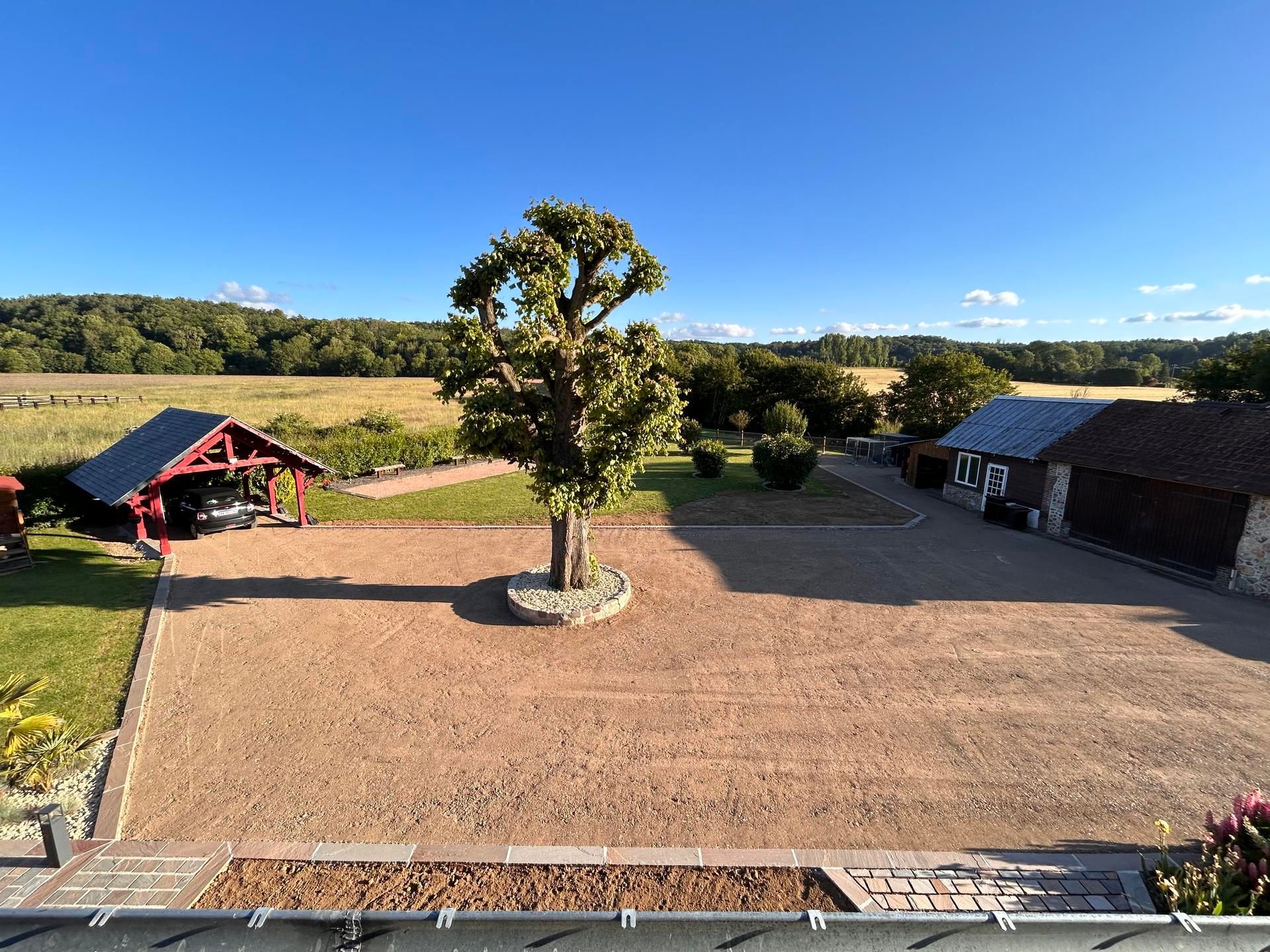 Cour ensoleillée avec un arbre central, un terrain gravillonné et des bâtiments aux toits rouges bordant des champs ouverts.