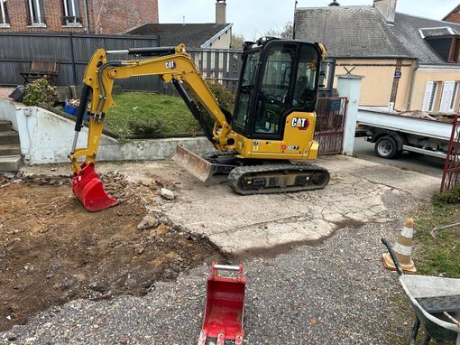Une minipelle jaune avec un godet rouge se trouve sur une allée de gravier près de maisons et d'un camion blanc.